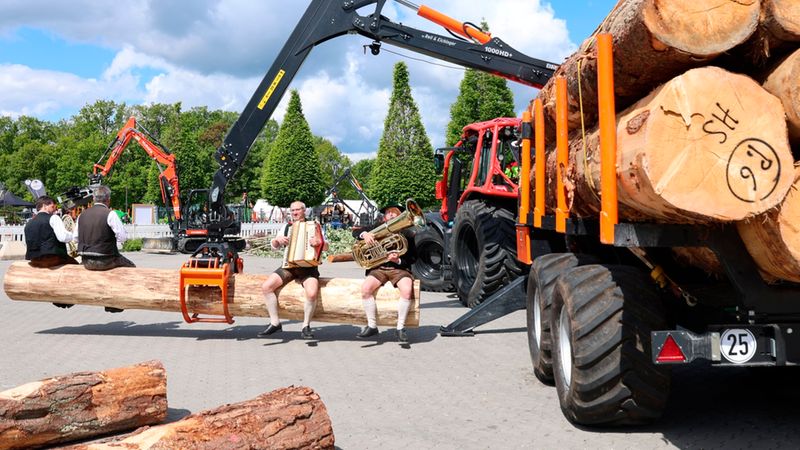 Eine Gruppe traditioneller Musikanten sitzt auf einem großen Holzstamm, der von einem Forstkran gehalten wird. Im Hintergrund sind weitere Maschinen und Bäume zu sehen.