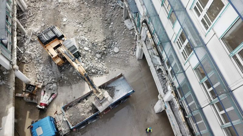 Ein Bagger lädt Schutt auf einen LKW auf einer Baustelle neben einem Gebäude.