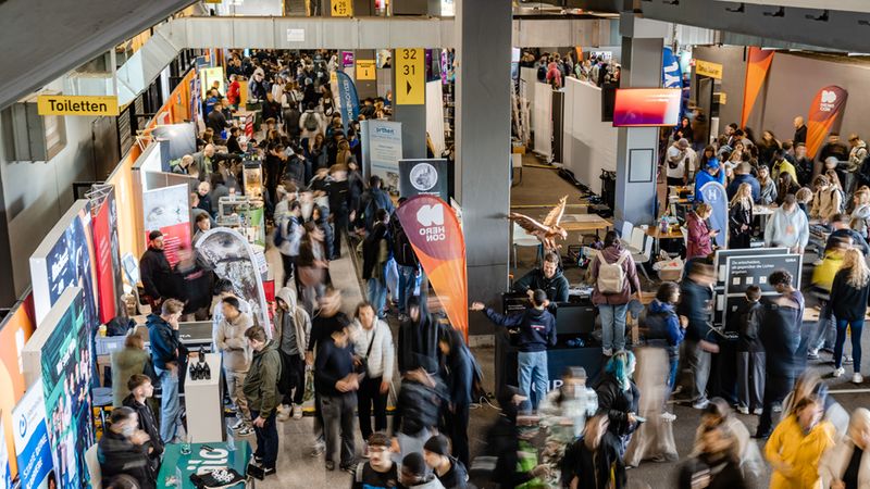Vogelperspektive einer belebten Messehalle mit zahlreichen Messeständen und vielen Besuchern, die zwischen Informationsständen und Präsentationen unterwegs sind; Bewegung und Unschärfe vermitteln die geschäftige Atmosphäre.