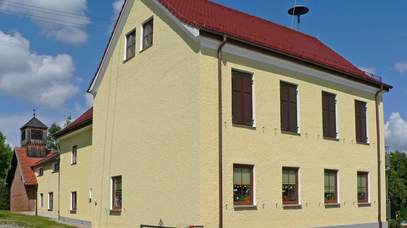 Hauptmotiv: ein gelbes zweistöckiges Dorfhaus mit rotem Ziegeldach, geschlossenen Fensterläden, kleinem Vorgarten und Zaun; im Hintergrund ein hölzerner Glockenturm, davor ein roter Hydrant und eine ruhige Straße bei blauem Himmel.