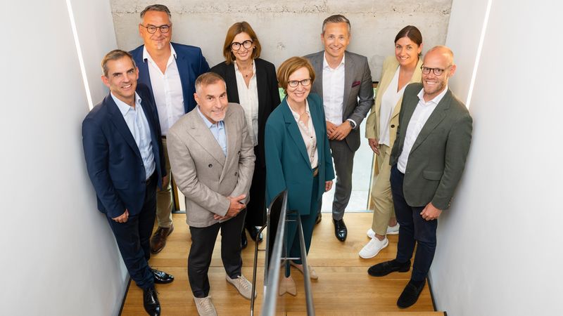 Gruppenporträt eines professionellen Geschäftsteams auf einer Treppe in einem modernen Innenraum. Acht Personen in Businesskleidung stehen dicht beieinander, blicken zur Kamera; helle Wände und Holzboden betonen die zeitgemäße, formelle Atmosphäre.