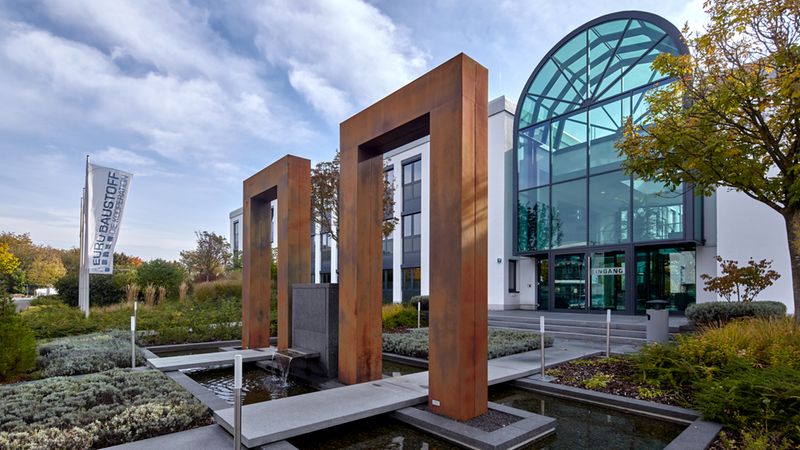 Vordergrund: ein rechteckiges Wasserbecken mit drei hohen Cortenstahl-Portalen und kleinem Wasserfall, durchzogen von Betonstegen. Im Hintergrund ein modernes Bürogebäude mit großer, glasüberdachter Eingangsfront, gepflegten Beeten und herbstlichen Bäumen sowie einer Firmenflagge am Rand.