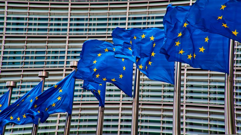 Mehrere wehende Flaggen der Europäischen Union (blau mit gelben Sternen) in einer Reihe vor der Fassade eines modernen Verwaltungsgebäudes; klare Farben, dynamische Bewegung durch Wind und eine geometrische Fensterstruktur im Hintergrund.