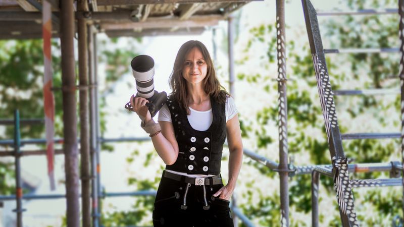 Porträt einer Fotografin mit großem Teleobjektiv, stehend auf einem Baugerüst. Die Frau trägt lässige Arbeitskleidung und hält die Kamera selbstbewusst in der Hand; der Hintergrund ist weichgezeichnetes Grün, natürliche Außenbeleuchtung.