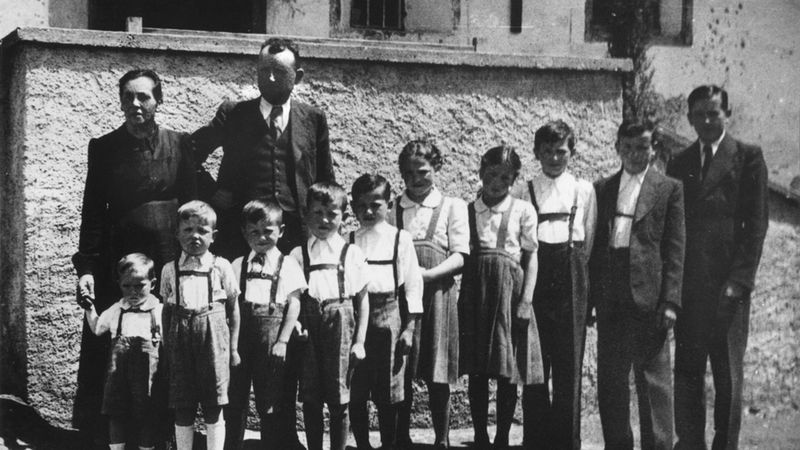 Schwarzweiß-Foto eines Familienporträts mit mehreren Kindern und Erwachsenen, aufgenommen im Freien vor einer Hausfassade. Die Personen stehen in einer Reihe; die Gesichter sind unkenntlich gemacht. Kleidung und Bildstil wirken historisch/vintage und vermitteln eine frühere Zeitperiode.