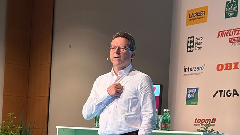 Ein Mann in weißem Hemd und dunklen Hosen steht auf einer grünen Bühne hinter einem Stehpult und hält einen Vortrag. Im Hintergrund sind Sponsorlogos an einer Konferenzwand sowie Pflanzen als Bühnen­deko zu sehen. Die abgebildete Person ist nicht identifizierbar.