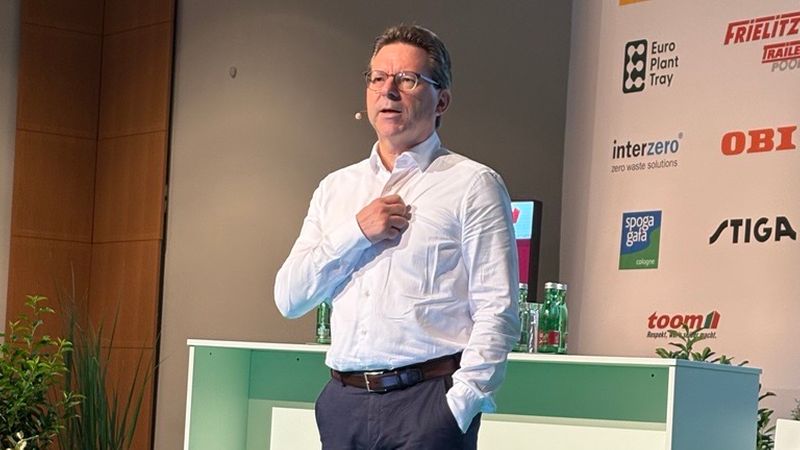 Ein Mann in weißem Hemd und dunklen Hosen steht auf einer grünen Bühne hinter einem Stehpult und hält einen Vortrag. Im Hintergrund sind Sponsorlogos an einer Konferenzwand sowie Pflanzen als Bühnen­deko zu sehen. Die abgebildete Person ist nicht identifizierbar.