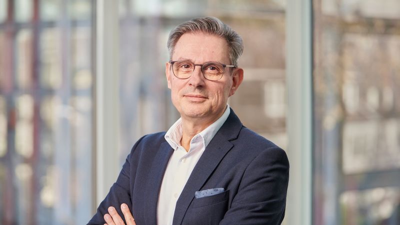 Ein Mann im Anzug steht mit verschränkten Armen vor einem Fenster.