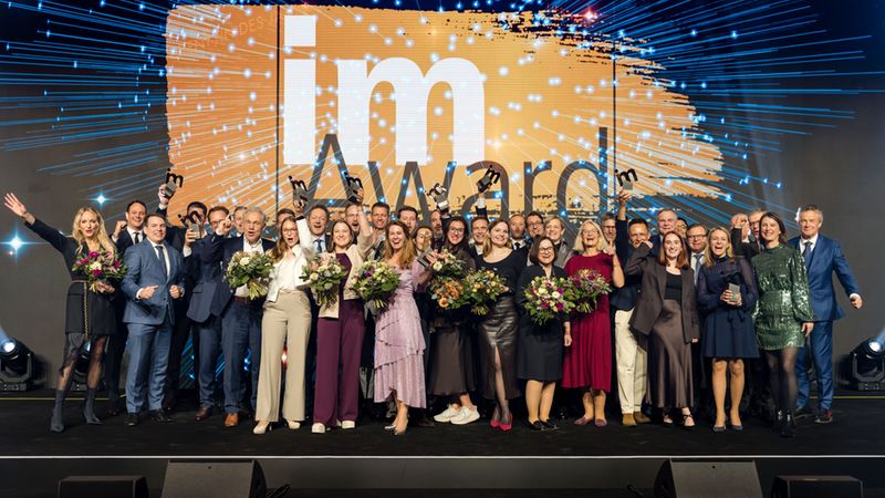 Großes Gruppenfoto von Preisträgern auf einer Bühne bei einer feierlichen Award-Verleihung; die Personen halten Trophäen und Blumensträuße, sitzen in festlicher Kleidung vor einer leuchtenden Medienwand.