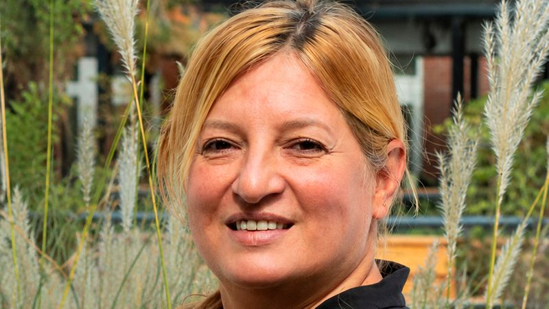 Porträt einer lächelnden, erwachsenen Frau mit blonden Haaren und schwarzem Oberteil, aufgenommen im Freien vor hohen Ziergräsern. Natürliche Beleuchtung, freundlicher Gesichtsausdruck und leichte Drehung des Oberkörpers zur Kamera, Umgebung wirkt wie ein Garten oder eine gestaltete Außenanlage.