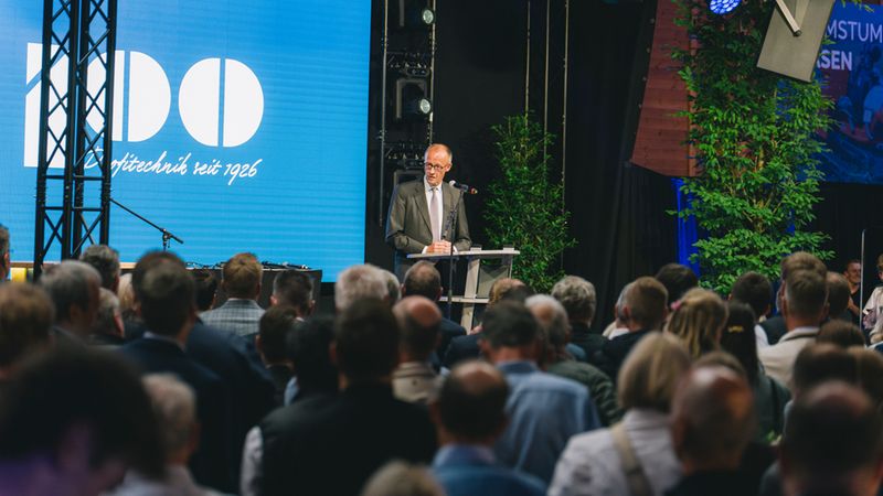 Hauptmotiv: Ein Redner am Podium hält eine Rede vor einem großen Publikum bei einer Firmenveranstaltung. Die Szene zeigt den präsentierenden Sprecher im Anzug, eine große LED-Leinwand mit Firmenlogo, Bühnenbeleuchtung, Mikrofon und Dekorationspflanzen. Aufnahme aus dem Publikum mit Blick auf Bühne und Redner.
