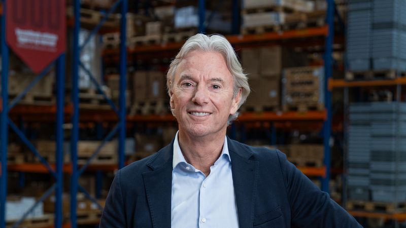 Ein lächelnder Geschäftsmann in Sakko und Hemd lehnt an einem roten Geländer in einer Lagerhalle. Im Hintergrund sind hohe Regale mit Kartons und Lagerbehältern zu sehen, die eine Logistik- und Versandumgebung zeigen. Professionelles Porträt eines Managers in einem industriellen Lager-Setting.