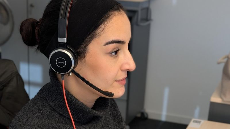Seitenprofil einer Person mit Headset, die an einem Schreibtisch sitzt; auf dem Tisch stehen eine Tasse und ein Notizbuch, die Person ist aufmerksam und arbeitet am Computer.
