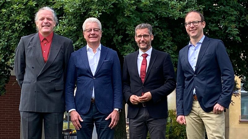 Hauptmotiv: Vier Männer in Anzügen stehen draußen vor einer dichten Heckenbepflanzung und posieren lächelnd für ein geschäftliches Teamfoto. Die Aufnahme zeigt formelle Kleidung, freundliche Körperhaltung und eine ruhige Außenatmosphäre.