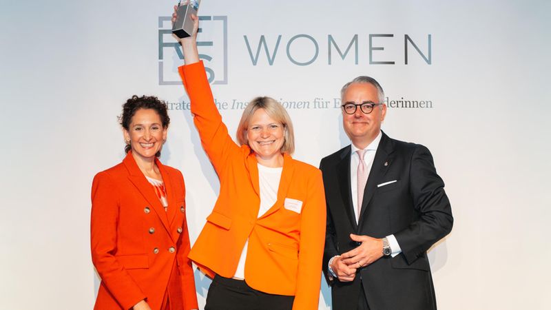 Hauptmotiv: Eine Frau in orangefarbenem Blazer hält jubelnd einen Pokal über dem Kopf. Sie steht auf einer Bühne vor einem Schriftzug "WOMEN" und wird von zwei Personen flankiert — einer Frau im ebenfalls orangen Anzug und einem Mann im dunklen Anzug. Die Szene zeigt Freude und Anerkennung bei einer Preisverleihung.