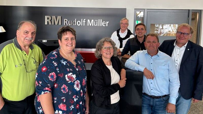 Hauptmotiv: Eine Gruppe von sieben Erwachsenen steht lächelnd vor einer Empfangstheke; im Hintergrund ist deutlich das Firmenlogo "RM Rudolf Müller" zu sehen. Die Szene vermittelt eine freundliche, informelle Geschäftsatmosphäre in einer Büro- oder Empfangsumgebung.