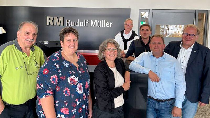 Hauptmotiv: Eine Gruppe von sieben Erwachsenen steht lächelnd vor einer Empfangstheke; im Hintergrund ist deutlich das Firmenlogo "RM Rudolf Müller" zu sehen. Die Szene vermittelt eine freundliche, informelle Geschäftsatmosphäre in einer Büro- oder Empfangsumgebung.