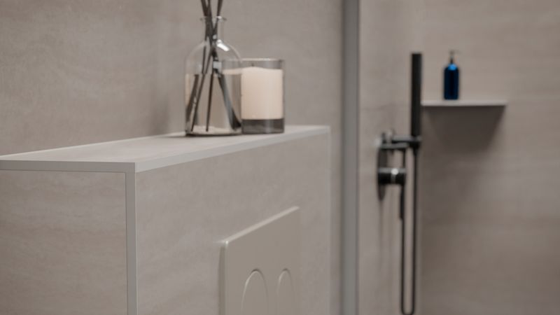Hauptmotiv: Nahaufnahme einer WC-Spülplatte neben einer schwarzen Duscharmatur in einem modernen, beige gehaltenen Badezimmer. Auf der Ablage sind eine Duftkerze und ein Glas mit Diffuser-Stäbchen zu sehen, im Hintergrund ein blauer Seifenspender.