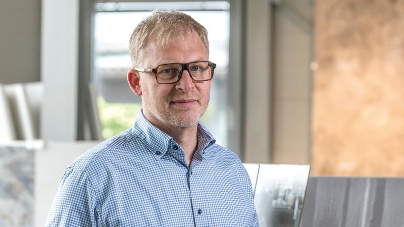 Porträt eines Mannes mit Brille in einem hellen, modernen Arbeitsumfeld. Er trägt ein kariertes Hemd und blickt leicht lächelnd in die Kamera.