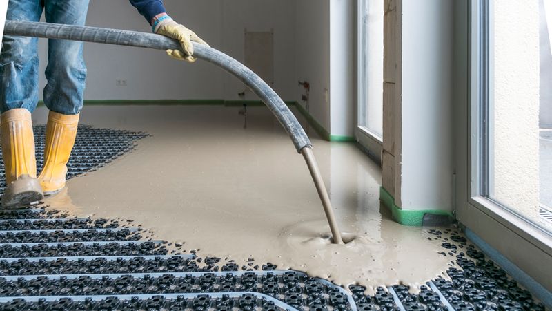 Arbeiter gießt Fließestrich mit einem Schlauch über eine installierte Fußbodenheizung; im Bild sind die Heizungsrohre in Noppenplatten, der flüssige Estrich, Schutzstiefel und ein Innenraum im Rohbau zu sehen.