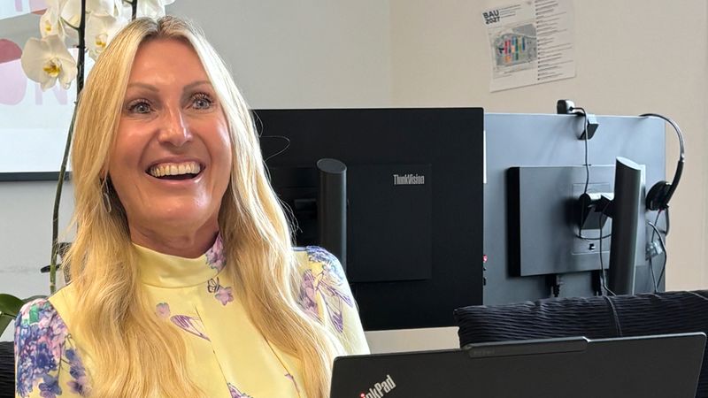 Eine lächelnde Frau mit langem blonden Haar sitzt an einem Schreibtisch und arbeitet an einem schwarzen Lenovo ThinkPad-Laptop in einem modernen Büro. Im Hintergrund sind zusätzliche Bildschirme, Headsets und eine Orchidee zu sehen. Die Szene vermittelt eine freundliche, professionelle Arbeitsatmosphäre.