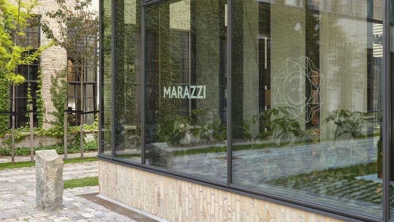 Glasfassade eines Showrooms mit deutlich sichtbarem MARAZZI-Logo auf der Fensterfront, reflektierenden Scheiben, Pflanzen im Innenraum sowie Kopfsteinpflaster und Backsteinmauer im Hof.