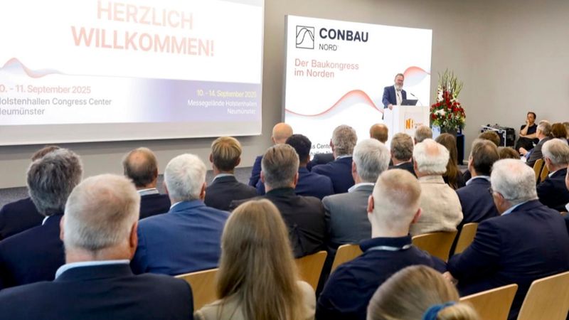 Ein Redner an einem Podium hält einen Vortrag beim CONBAU NORD Baukongress vor einem sitzenden Publikum in einem Konferenzraum; im Hintergrund ist ein Willkommens-Screen und Konferenzbranding sichtbar, die Atmosphäre wirkt professionell und geschäftlich.