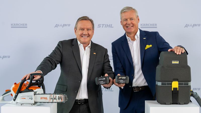 Zwei Männer in Anzügen präsentieren akkubetriebene Geräte vor einem Messestand mit Hersteller-Logos: eine STIHL Akku-Kettensäge, zwei Akkupacks und ein KÄRCHER Sauger. Professionelle Produktvorstellung in klarer, werblicher Inszenierung.
