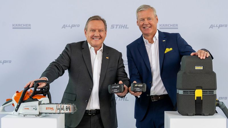 Zwei Männer in Anzügen präsentieren auf einem Messestand eine STIHL Akku-Kettensäge, dazugehörige Akkus und einen KÄRCHER Industriesauger; Logos und Produktdetails sind sichtbar, die Szene wirkt wie eine Produktvorstellung.