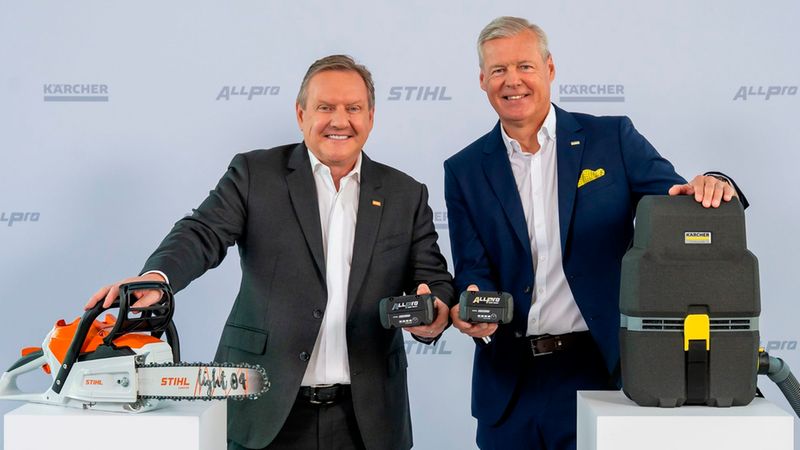 Zwei Männer in Anzügen präsentieren auf einem Messestand eine STIHL Akku-Kettensäge, dazugehörige Akkus und einen KÄRCHER Industriesauger; Logos und Produktdetails sind sichtbar, die Szene wirkt wie eine Produktvorstellung.