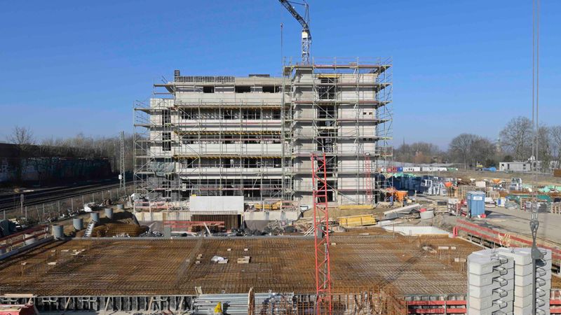 Mehrere Stockwerke eines Wohn- oder Bürogebäudes im Rohbau sind von Gerüsten umgeben, ein hoher Baukran ragt über die Baustelle; im Vordergrund sichtbare Bewehrung, Betonflächen und Baumaterialien auf einem weitläufigen Baugelände unter klarem blauem Himmel.