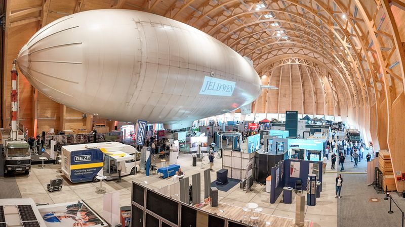 Ein großes weißes Luftschiff (Zeppelin) hängt über zahlreichen Messeständen in einer weiten Ausstellungshalle mit hohem hölzernem Gewölbe. Besucher schlendern zwischen Ständen, Fahrzeuge und Messeausrüstung sind sichtbar, die Szene wirkt wie eine Industrie- oder Technologiemesse in einem markanten Holzbau.