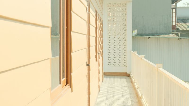 Ein schmaler, sonnendurchfluteter Balkonflur entlang einer pastellfarbenen Hausfassade mit Holzfenstern, einem weißen Geländer und markanten Schattenmustern auf dem Boden.