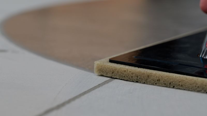 Nahaufnahme eines Fugenschwamms mit Schaumstoffunterlage auf einer hellen Fliesenoberfläche, sichtbar ist eine Fugenlinie und ein Teil eines Werkzeugs/Griffs; Szene vermittelt Fliesenverfugung und Reinigung beim Handwerk.