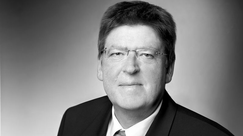 Schwarz-Weiß-Studioporträt eines älteren Mannes in Anzug und Krawatte mit Brille. Kopf- und Schulteraufnahme vor neutralem Hintergrund, seriöser und professioneller Ausdruck. Hauptmotiv: Porträt eines Mannes.