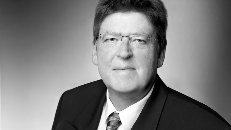 Schwarzweiß-Business-Porträt eines erwachsenen Mannes mit Brille, Anzug und Krawatte vor neutralem Hintergrund. Frontalaufnahme mit weichem Licht, konzentriert auf Gesicht und obere Brustpartie. Hauptmotiv: männliches Porträt.