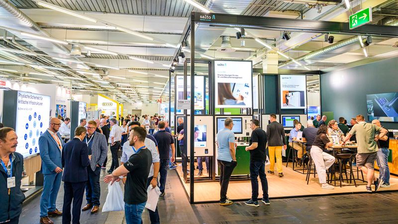 Belebte Messehalle mit mehreren Ausstellungsständen, Besuchern, Informationsdisplays und moderner Beleuchtung. Personen führen Gespräche, betrachten Produktpräsentationen und interagieren an Messeständen; Fokus auf Messeumgebung, Networking und Technologiepräsentationen.