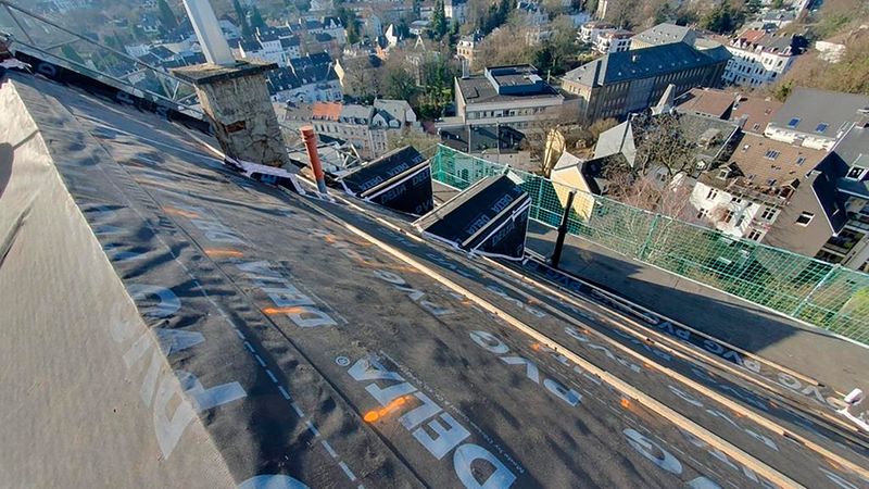 Blick von einem steilen Dach während einer Dachsanierung: sichtbare DELTA-Unterspannbahn auf der Dachfläche, Holzlatten, Dachanschlüsse am Schornstein sowie Baustellensicherung mit Gerüst. Im Hintergrund ist eine Stadtansicht mit Wohnhäusern und bewaldeten Hügeln zu sehen.