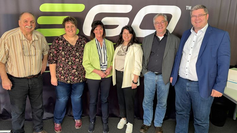 Sechs Personen stehen nebeneinander und lächeln vor einem dunklen Hintergrund mit großem EGO-Firmenlogo. Die Gruppe trägt business-casual Kleidung und wirkt entspannt; das Foto wurde bei einer Indoor-Veranstaltung oder einem Meeting aufgenommen. Keine individuellen Personen identifiziert.