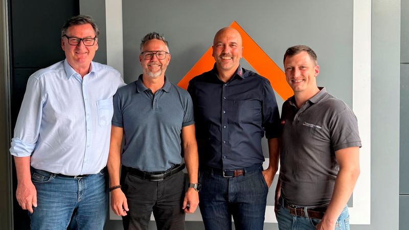 Gruppenfoto mit vier Männern in lässiger Business-Kleidung, die nebeneinander vor einer Wand mit einem orangefarbenen, aufwärts gerichteten Firmenlogo stehen. Heller Rahmen und graue Wand im Hintergrund, entspannte Haltung und freundliche Mimik, geeignet als Team- oder Firmenbild.
