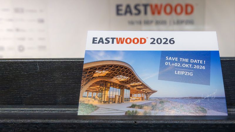 Fotografie einer Save-the-Date Karte für "EASTWOOD 2026" auf einem dunklen Holzregal. Die Karte zeigt prominent den Schriftzug "EASTWOOD 2026", das Datum "01.+02. Okt. 2026" und den Ort "Leipzig" sowie ein Bild einer modernen Holz-Architektur oder Pavillon-ähnlichen Halle. Das Motiv kombiniert Veranstaltungshinweis und architekturbezogenes Foto in einer Werbekarte.