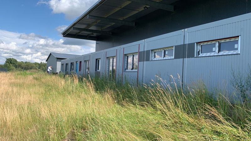 Industriehalle mit hohem Gras im Vordergrund. Das langgestreckte Gebäude zeigt eine graue Metallfassade mit schmalen Fenstern; der Vordergrund ist von verwildertem, hohem Gras geprägt. Am linken Bildrand ist eine Person im Hintergrund zu sehen, darüber ein blauer Himmel mit Wolken.