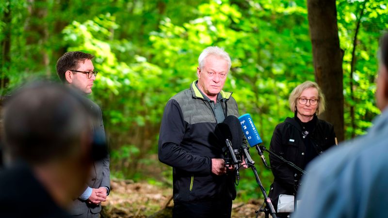 Außenaufnahme einer Pressekonferenz im Wald: Im Vordergrund steht Bundesminister Alois Rainer als Hauptmotiv und spricht vor mehreren Mikrofonen, begleitet von zwei Personen im Hintergrund. Unscharfe Zuschauer sind am Bildrand erkennbar, die Szene ist von grüner Baum- und Blattkulisse umgeben und vermittelt einen seriösen, informativen Charakter.