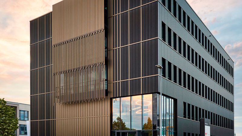 Modernes mehrstöckiges Bürogebäude als Hauptmotiv: dunkle vertikale Metallfassade mit Fassadendetails, große Glasfront am Eingangsbereich, Treppenstufen und Umfeld in einer urbanen Bürolandschaft, aufgenommen bei warmem Sonnenuntergangslicht.