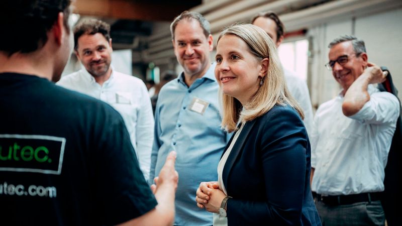 Eine lächelnde Geschäftsfrau in einem dunkelblauen Blazer führt auf einer Firmenveranstaltung ein Gespräch mit mehreren Kollegen; die Szene vermittelt eine professionelle, freundliche Atmosphäre mit aktivem Networking und Austausch.