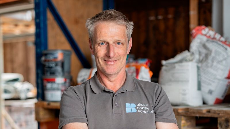 Porträt eines lächelnden Handwerkers in Arbeitskleidung, der im Lager vor Regalen mit Baustoffen steht und einen Fliesenschneider hält. Die Szene zeigt ein organisiertes Lagermilieu mit Säcken und Paletten voller Material, typische Arbeitsumgebung eines Handwerksbetriebs.