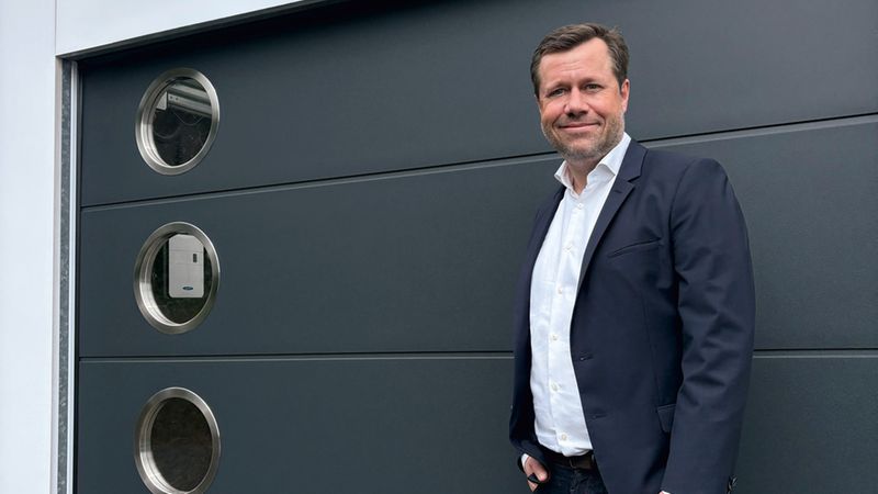 Eine Person in Businesskleidung steht lächelnd vor einem dunklen Novoferm-Garagentor mit ovalem Firmenlogo oberhalb und vier runden Fensteröffnungen, Außenaufnahme bei Tageslicht.