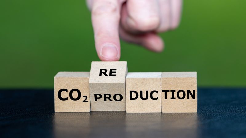 Holzbausteine mit der Aufschrift "CO2 pro duction", wobei eine Hand einen Würfel mit der Beschriftung "RE" daraufsetzt. Das Hauptmotiv CO2-Reduktion symbolisiert die Verringerung der CO2-Produktion und Klimaschutz vor unscharf grünem Hintergrund.