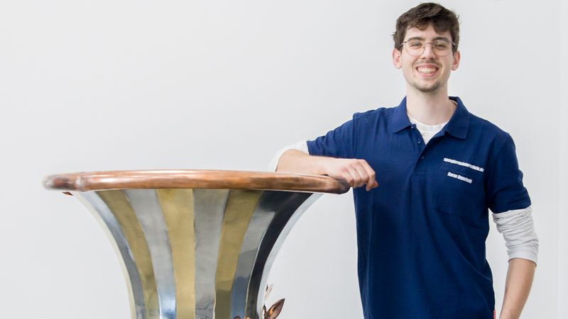 Eine große, bauchige Metallvase mit abwechselnden gold- und silberfarbenen Streifen und kupferfarbenen Rosenapplikationen steht vor einem neutralen weißen Hintergrund. Eine lächelnde Person in dunkler Arbeitskleidung lehnt locker an der Vase und vermittelt den Maßstab. Die Vase hat eine polierte, reflektierende Oberfläche und dekorative Kupferdetails am Fuß und in der Mitte.