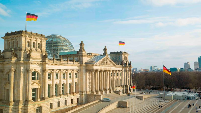 Luftaufnahme des Reichstagsgebäudes in Berlin als Hauptmotiv, mit der markanten Glaskuppel und mehreren wehenden deutschen Flaggen vor klarem blauem Himmel; breiter Platz und wenige Spaziergänger im Vordergrund.